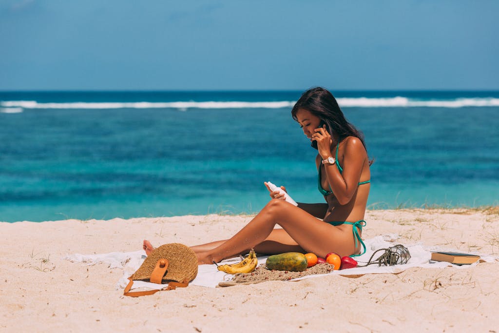 A woman in a bikini sits on the beach, enjoying a sunny day while talking on the phone and applying sunblock.