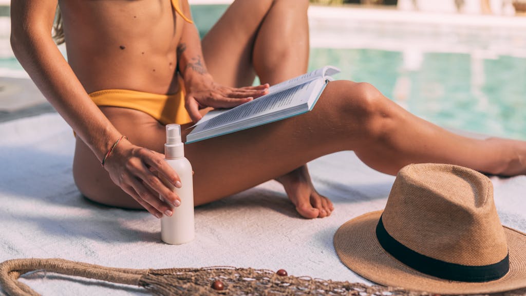 Woman in bikini reading by the pool with sunscreen and a hat, enjoying summer relaxation.