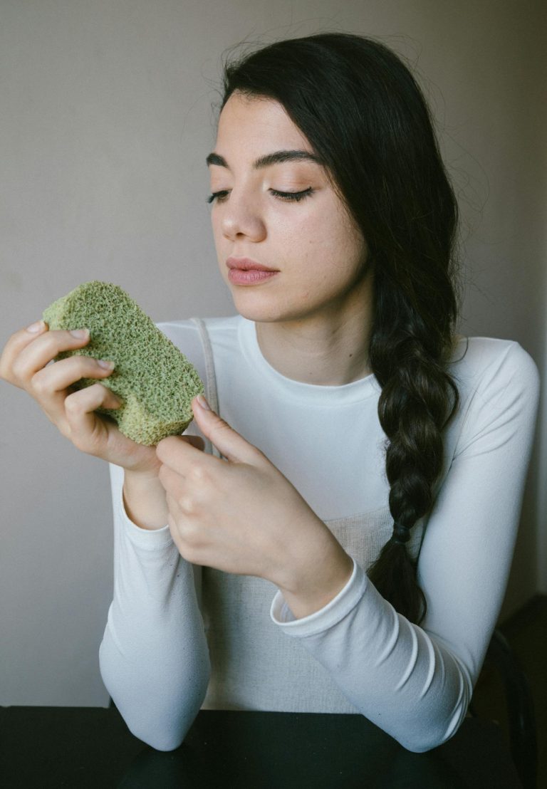 Young woman holding a green eco-friendly sponge indoors, promoting natural skincare.