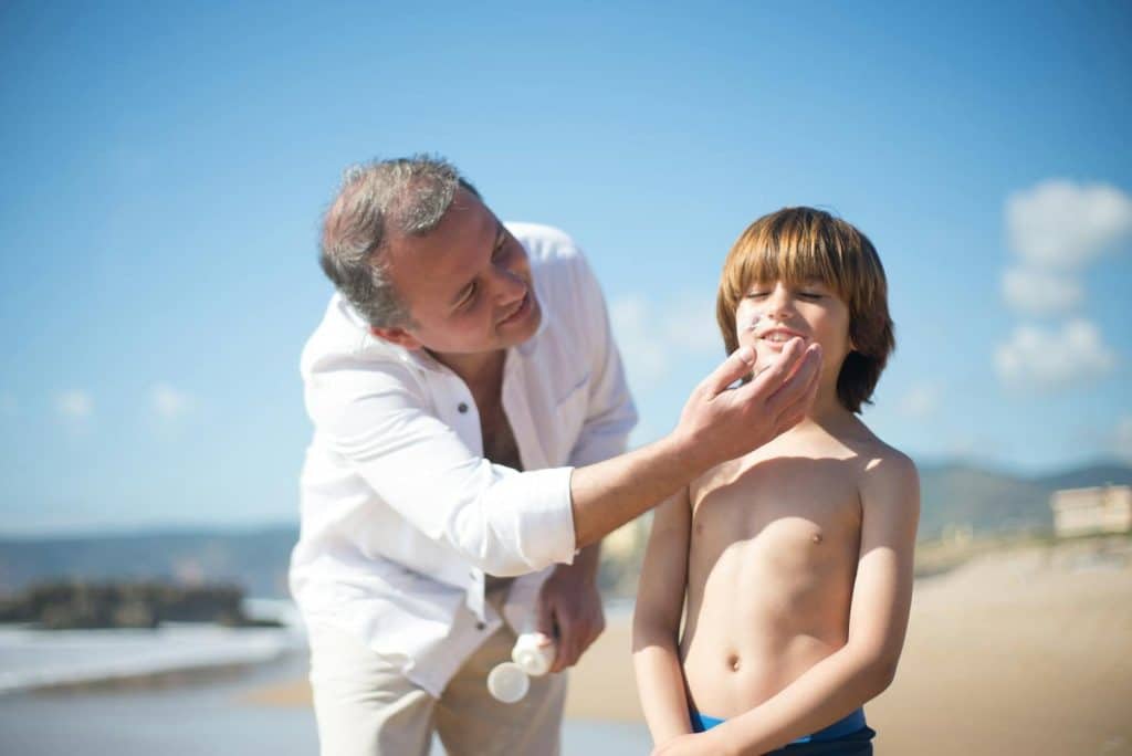 A father applies sunscreen to his smiling son on a sunny beach day.