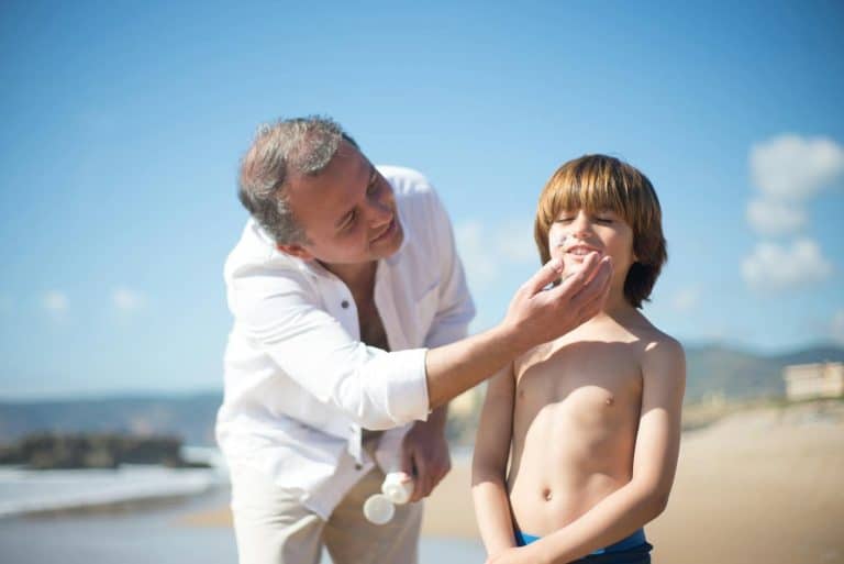 A father applies sunscreen to his smiling son on a sunny beach day.