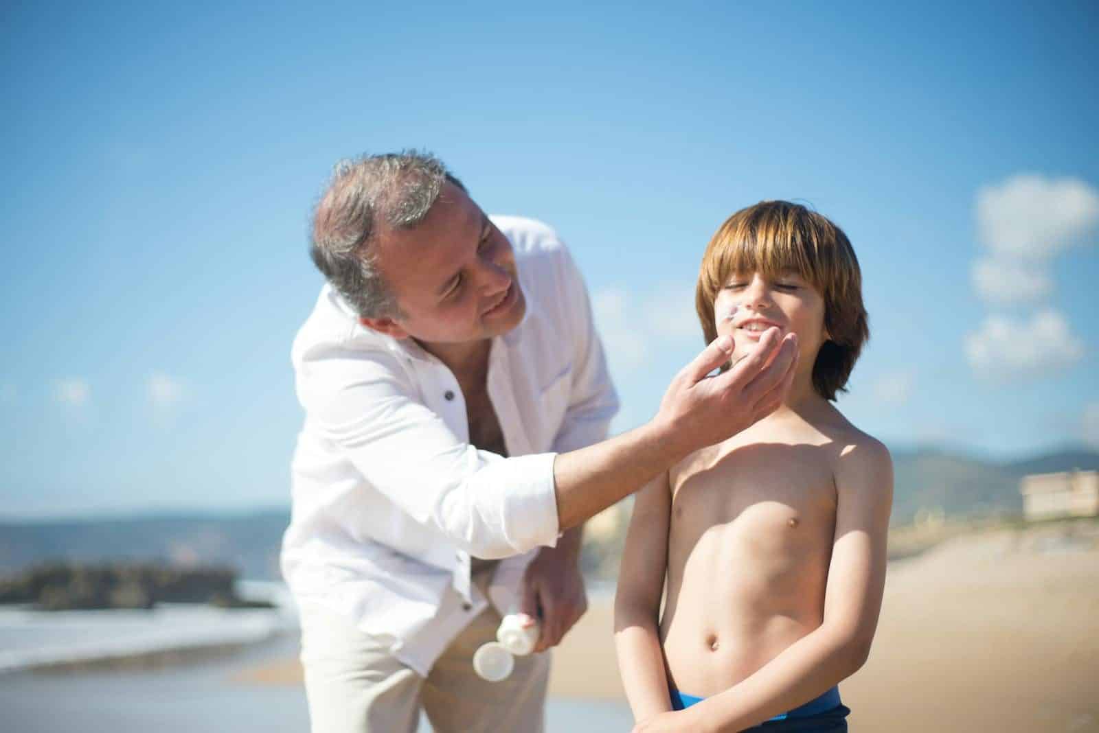 A father applies sunscreen to his smiling son on a sunny beach day.