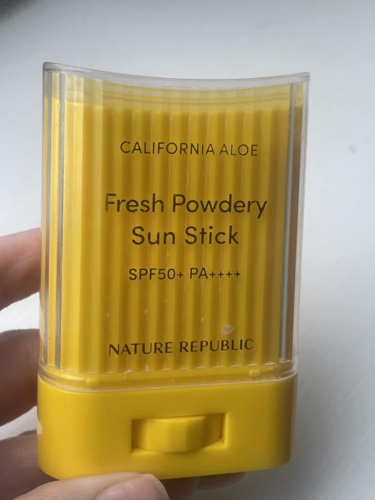 A hand holding a yellow Nature Republic California Aloe Fresh Powdery Sun Stick SPF 50+ inside its clear plastic protective cap against a plain white background.